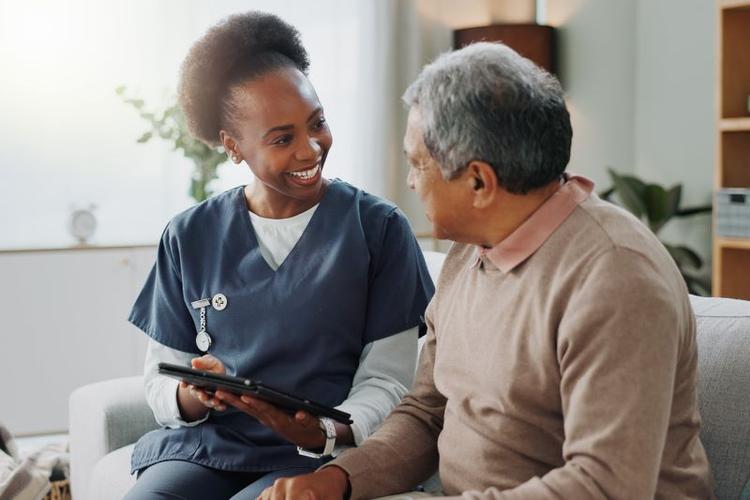 Nurse showing a tablet to an elderly man, smiling together in a living room setting.